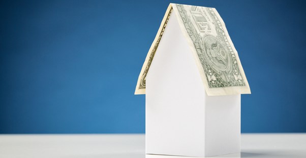 White model home with dollar bill on top as the roof in front of a navy blue background