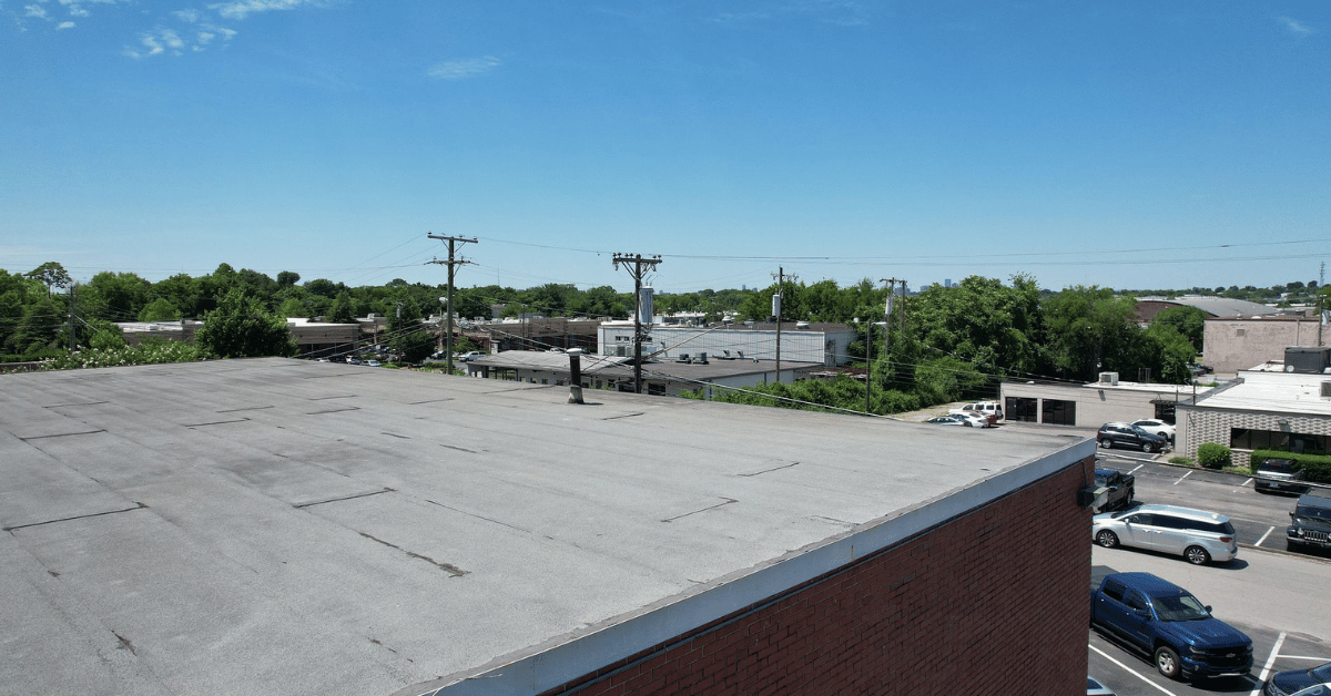 A flat commercial roof with visible seams, overlooking a business district with trees and power lines under a clear blue sky.