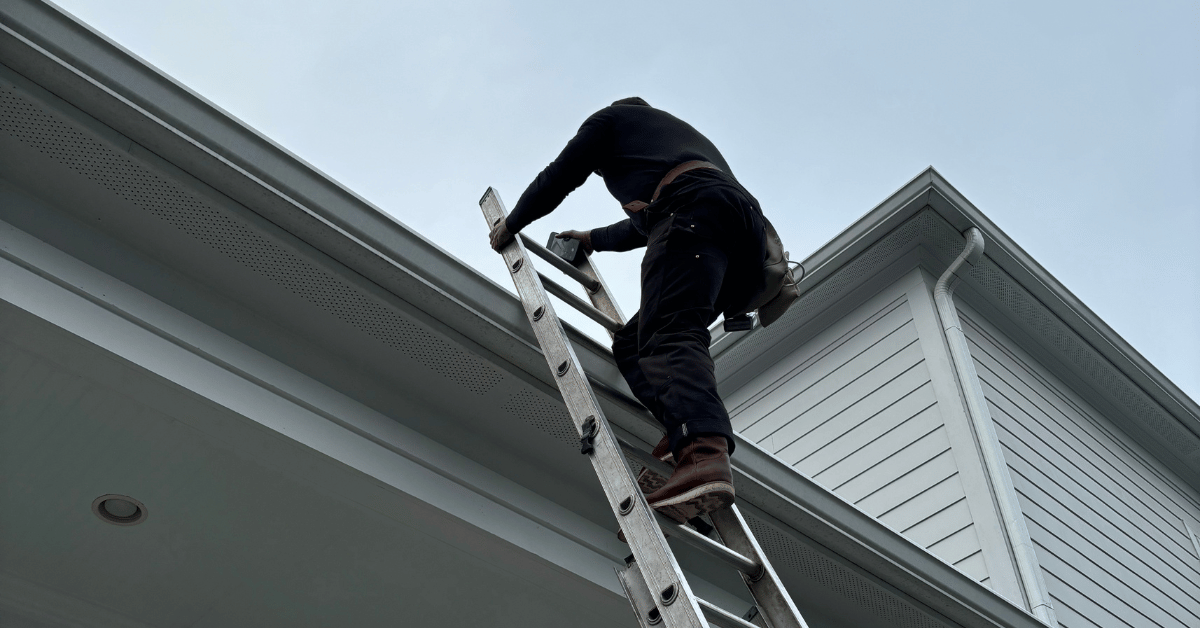 A roofer on a ladder cleaning gutters along the edge of a house roof.