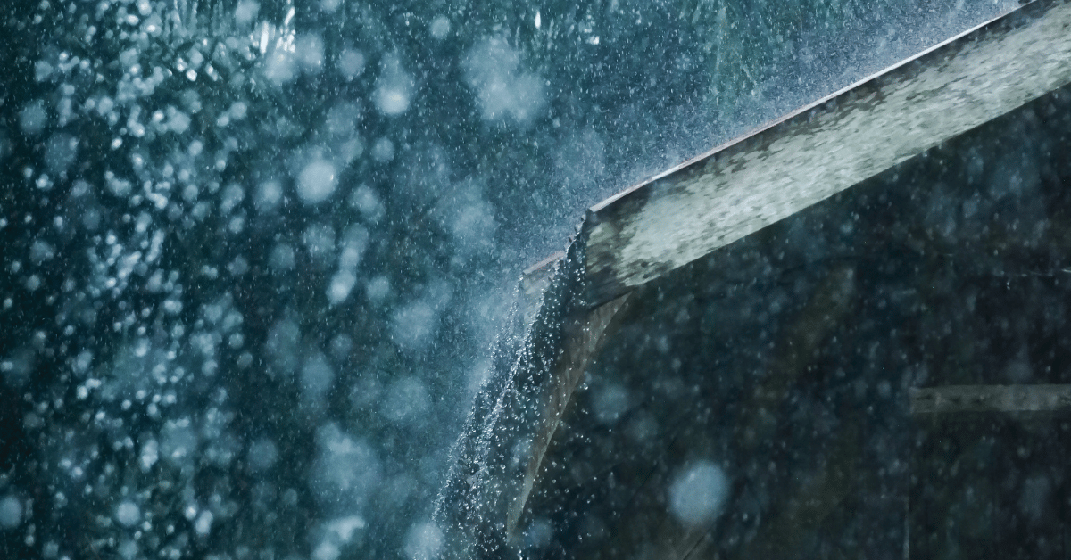 A moody shot of rain and mist cascading from the edge of a roof, creating an atmospheric view of storm conditions.