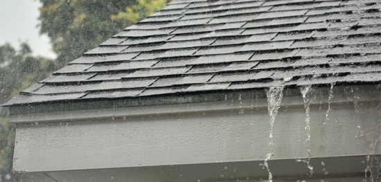 Rain heavily falling on a shingle roof and overflowing the gutters, indicating potential water damage or gutter blockage.