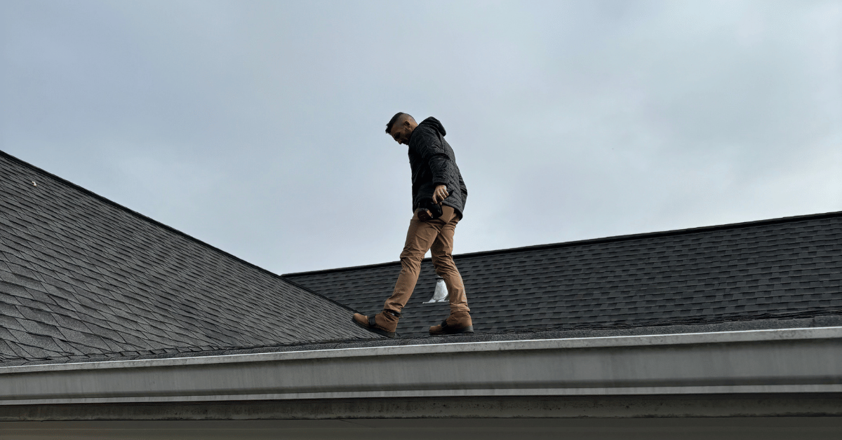 A roofer in a black jacket walking carefully on a sloped roof while carrying tools.