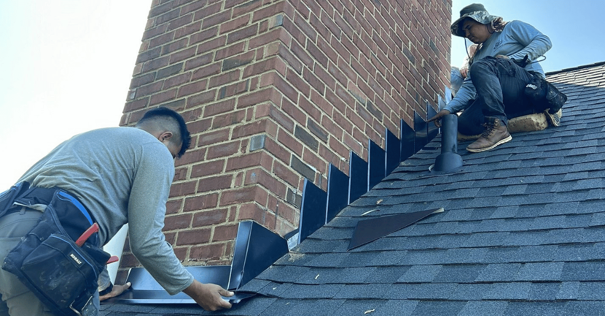 Two roofing professionals installing step flashing along a brick chimney.