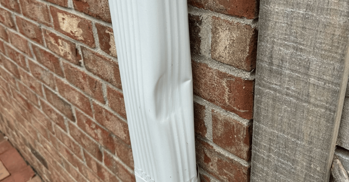 A white aluminum downspout with visible dents caused by hail.