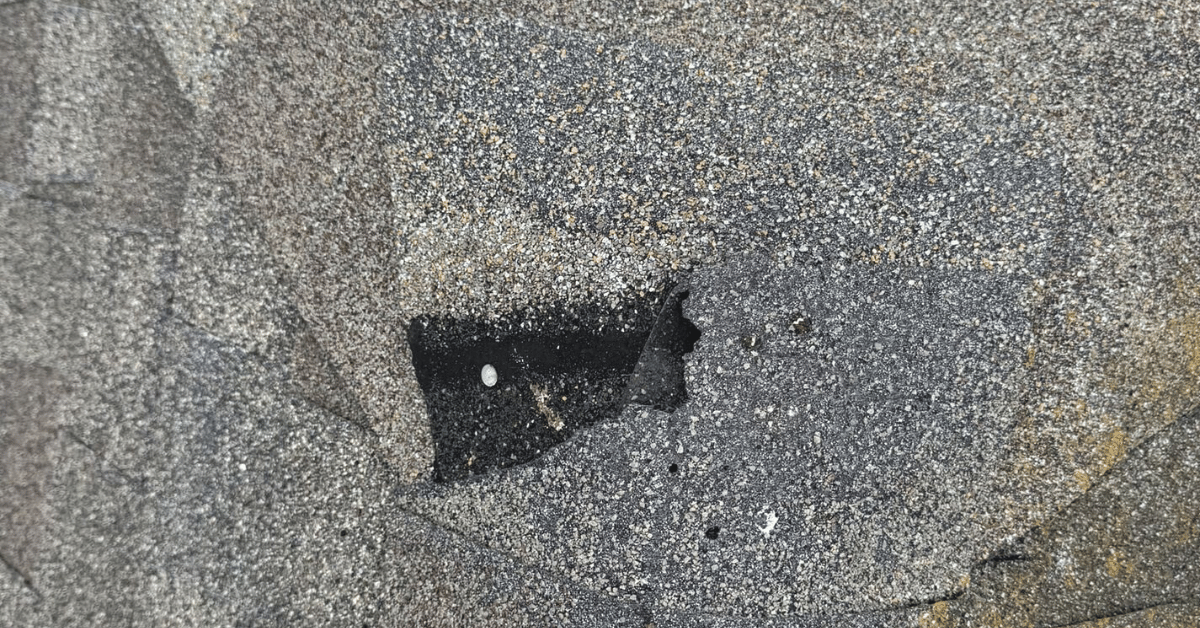 A damaged asphalt shingle with a hole exposing the black substrate underneath.