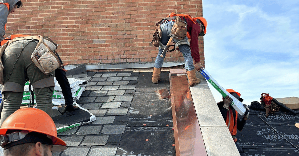 A roofing crew installing asphalt shingles and copper flashing on a brick building.