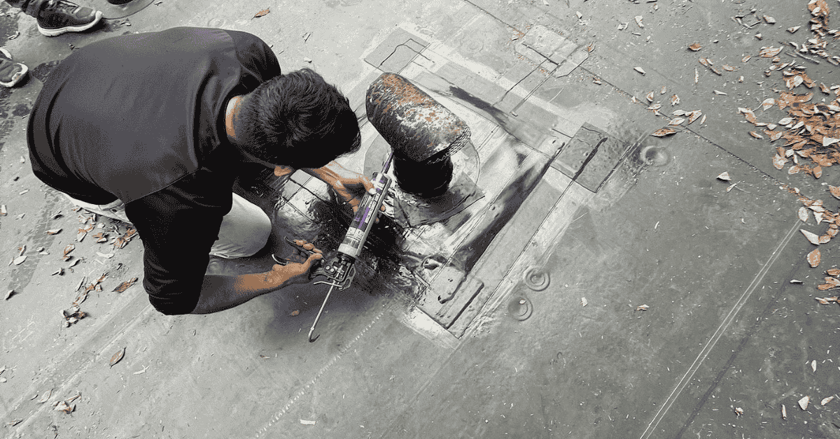 A roofer applying sealant around a damaged pipe boot on a flat roof.