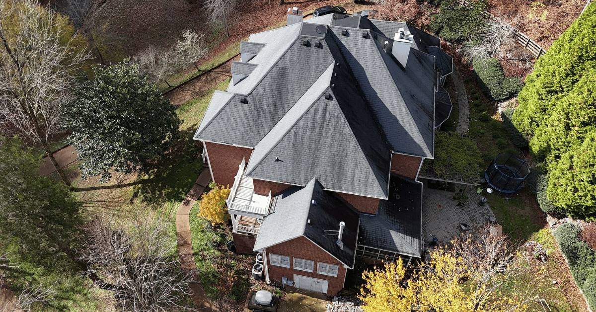 A drone shot capturing the roof of a large brick home surrounded by trees and landscaping.