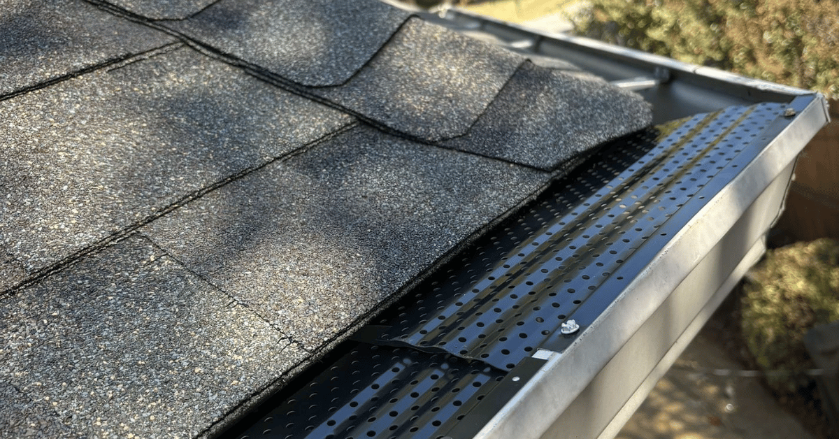 A section of a roof with lifted shingles near the gutter edge.