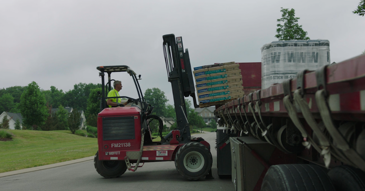 A worker operating a forklift to unload roofing materials from a truck in a suburban neighborhood.