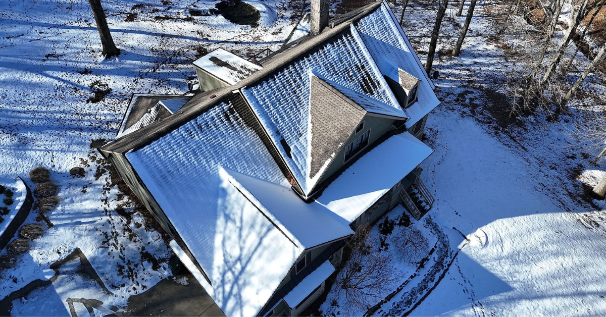 A large home with a steeply pitched roof covered in snow, surrounded by a wooded area.