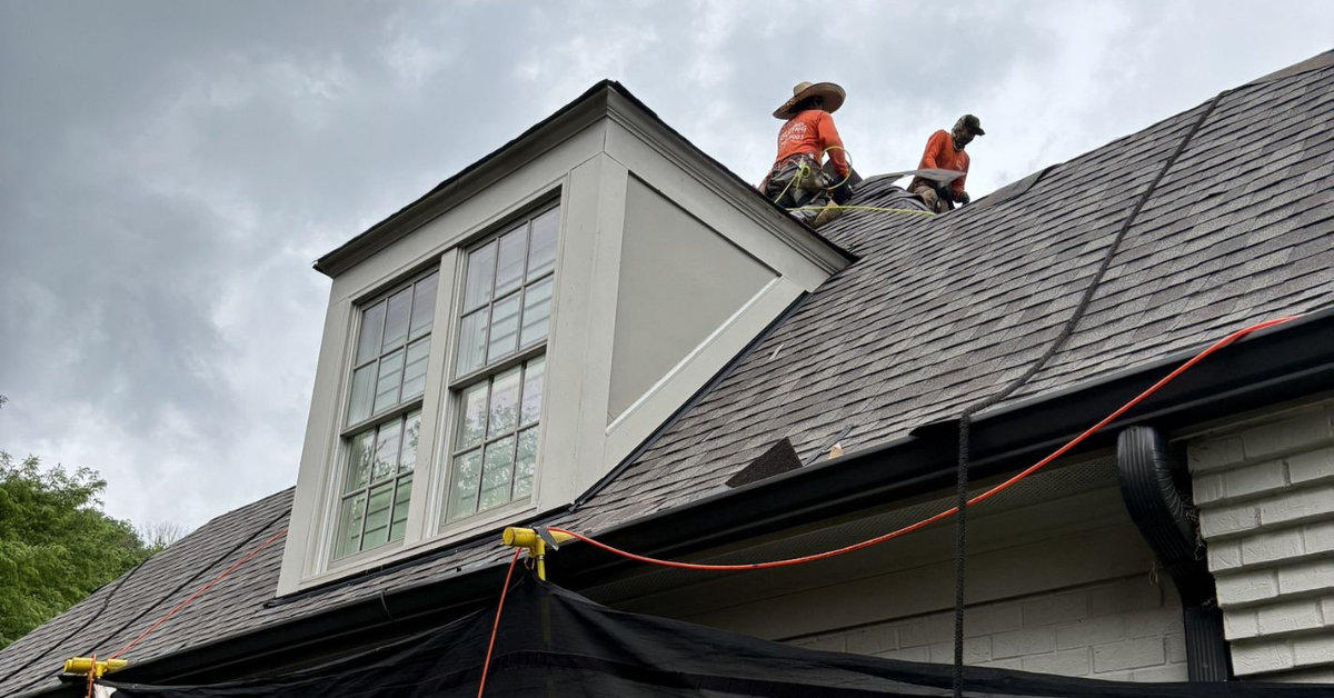 Two roofers in safety gear repairing a steep roof under cloudy skies, emphasizing the urgency of summer maintenance.