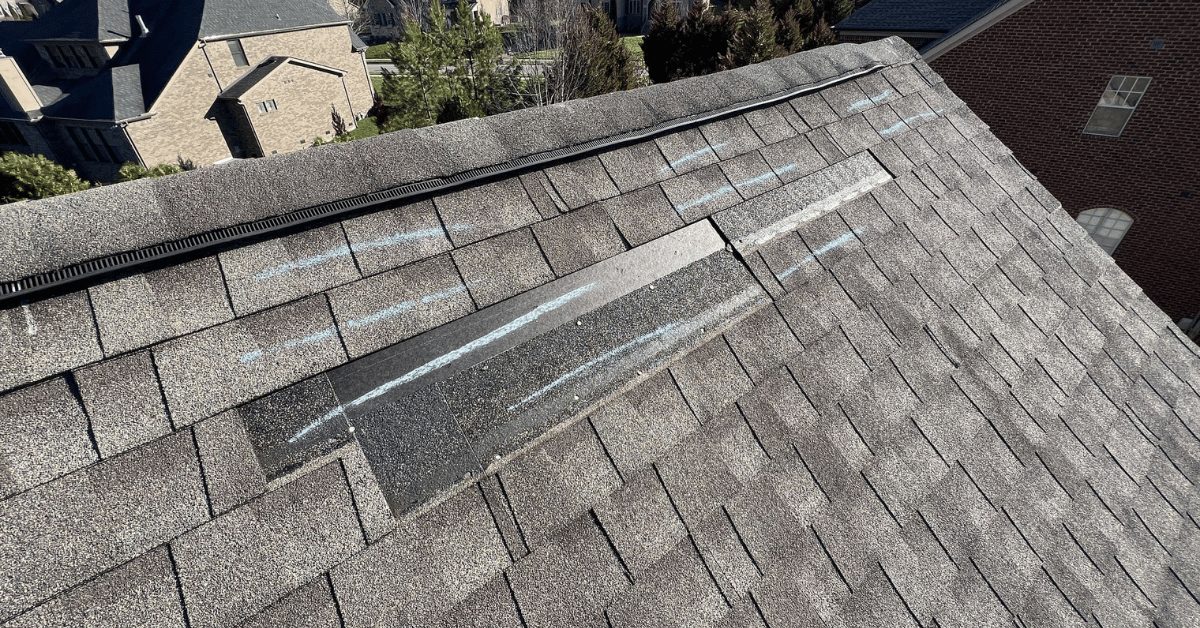Close-up of damaged shingles marked with chalk, highlighting wind impact on ridge cap during inspection.