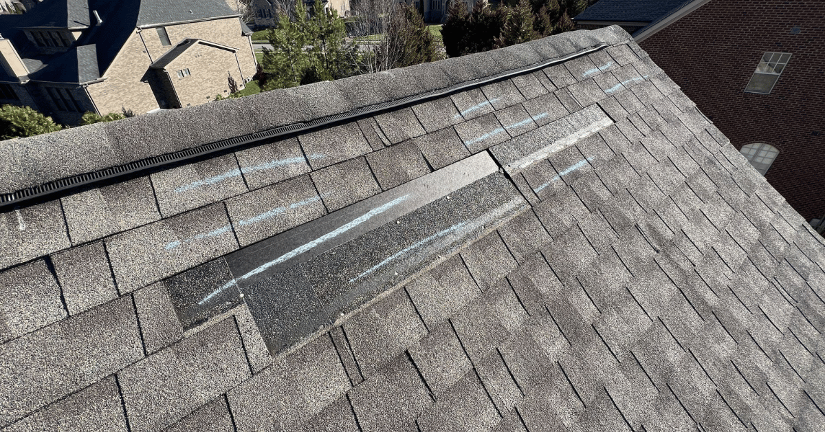 Wind-Damaged Shingles Identified During Roof Inspection Close-up of damaged shingles marked with chalk, highlighting wind impact on ridge cap during inspection.
