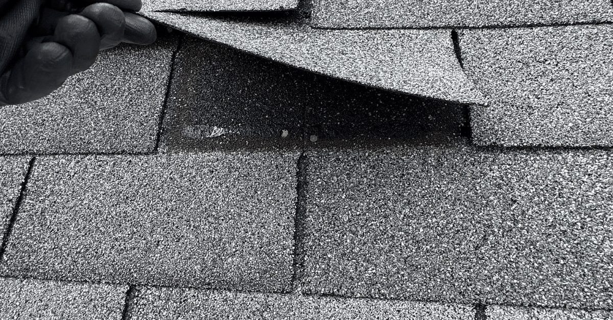 A roofer lifts an asphalt shingle to reveal proper nail placement and sealing during an inspection.