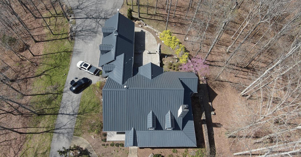 Aerial shot of a modern home with a dark gray standing seam metal roof surrounded by trees, illustrating a premium roofing option.