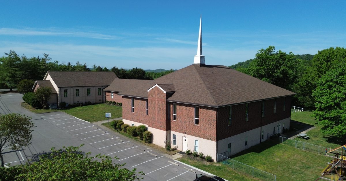 Ground-level drone view of West Harpeth Primitive Baptist Church with new brown shingles, white steeple, and brick exterior against a blue sky