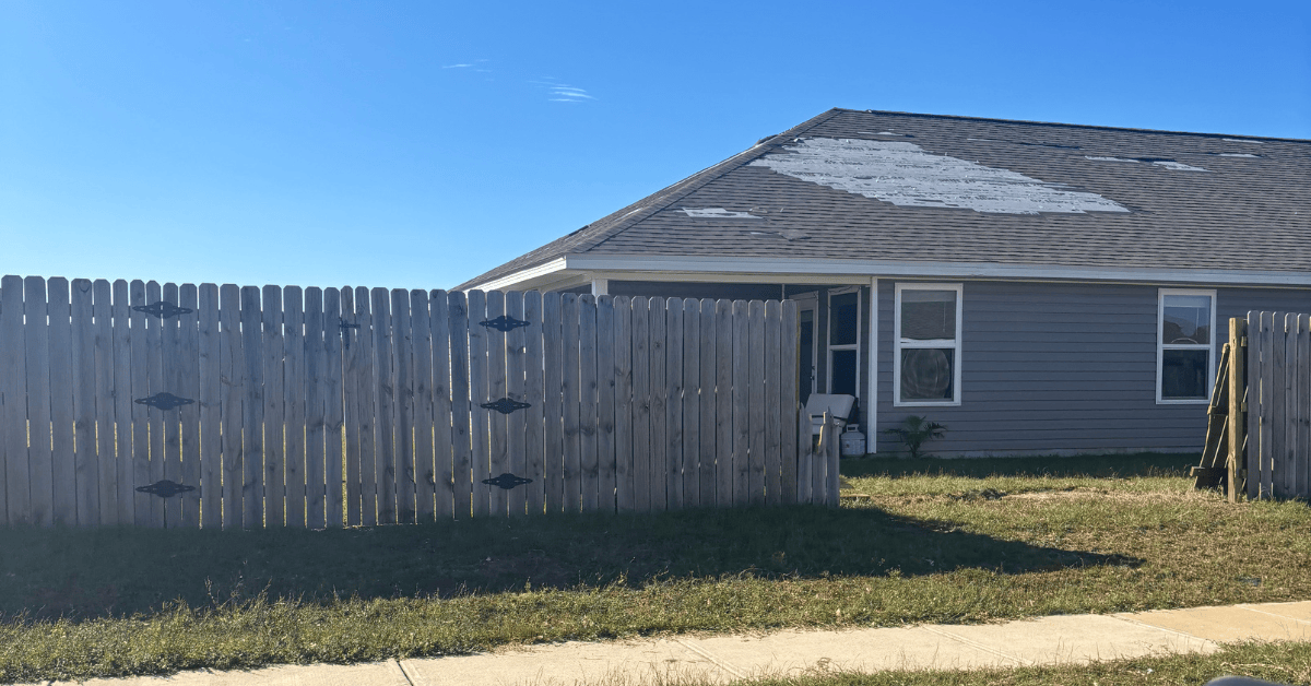 Gray shingle roof with a large section of shingles torn away, exposing white underlayment above a wood fence