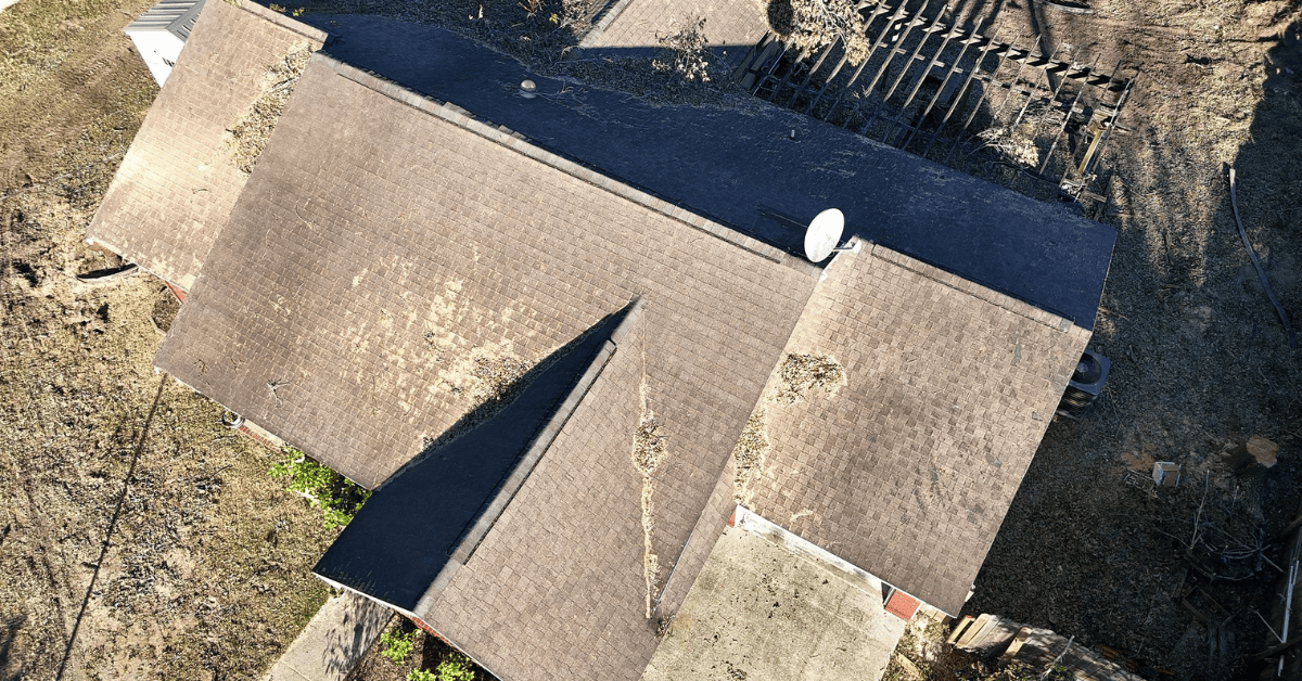 Aerial drone view of a brown asphalt shingle roof scattered with storm debris, with a broken wood fence in the yard