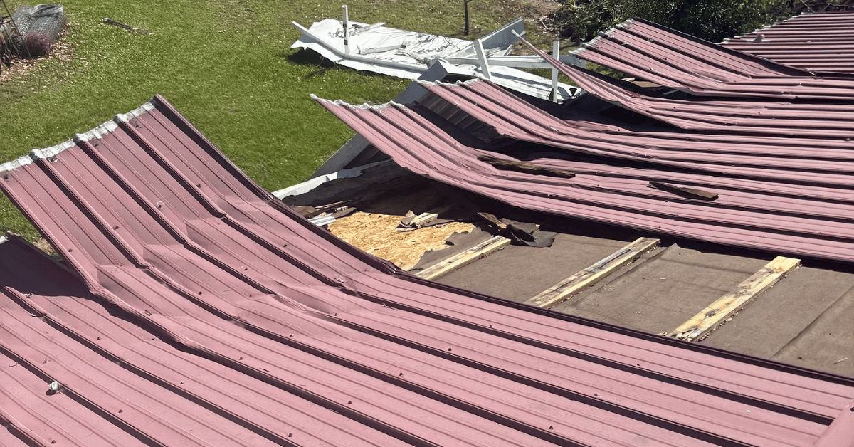 Burgundy metal roof panels peeled back by wind, exposing underlayment and roof decking, with torn panels in the yard